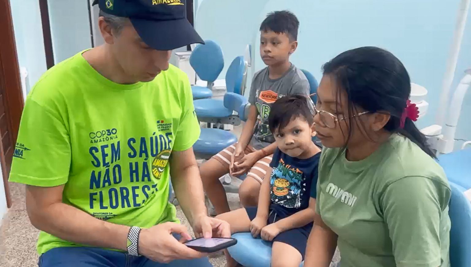 Fotografia registra um homem, com celular na mão, atendendo uma mulher, acompanhada de duas crianças.