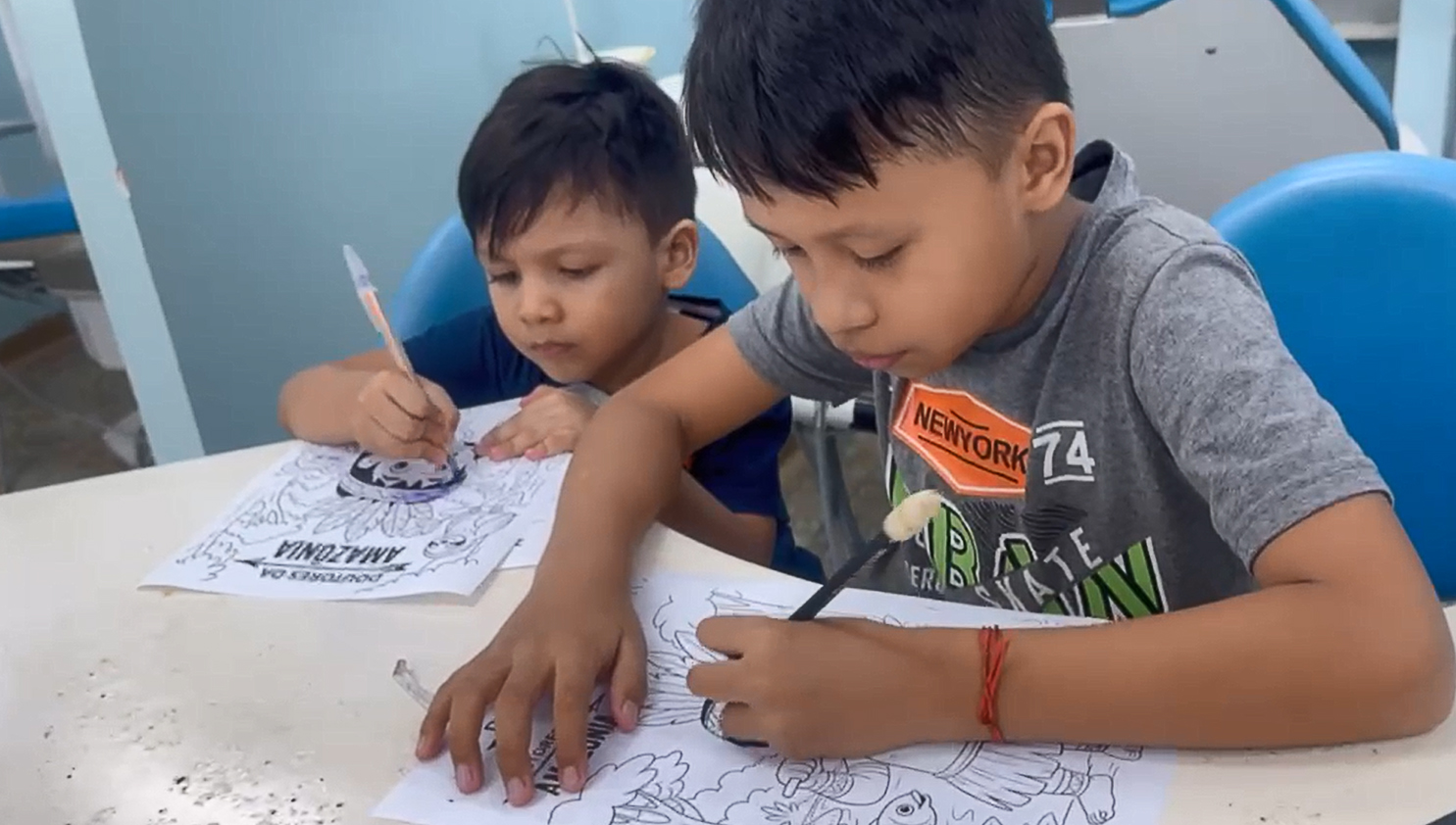 Fotografia registra duas crianças, de cabeças baixas, pintando desenhos em folhas de papel.